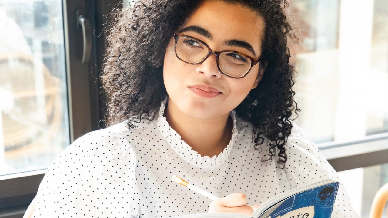 Garota negra pensativa, com livro e caneta na mão.
