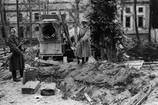 Dois soldados russos apontam para o que seria o túmulo de Hitler nos fundos do seu bunker em Berlim. Ao lado deles estão latas de gasolina. (Créditos: Fred Ramage/Keystone/Getty Images)