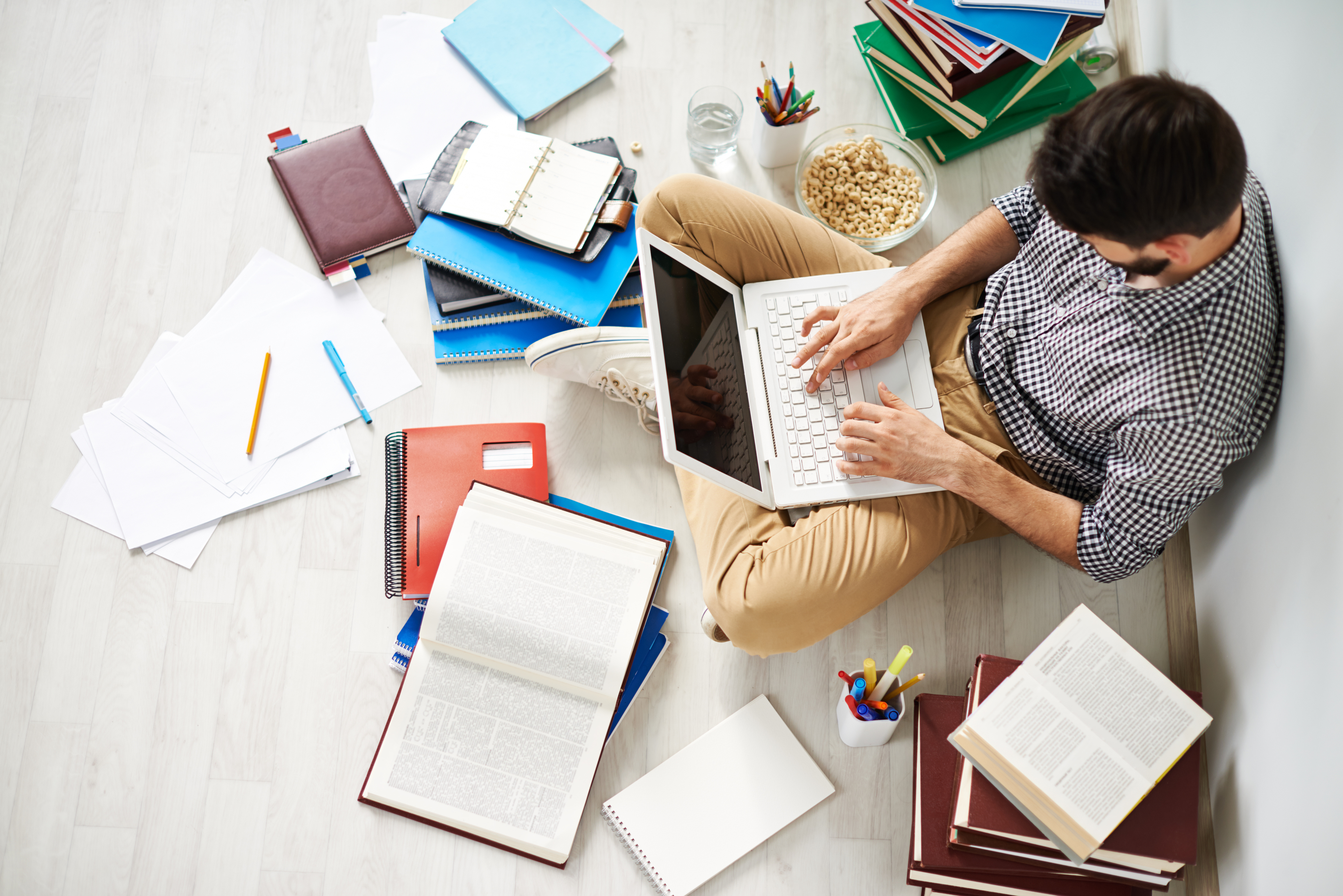 Jovem sentado no chão usando laptop, cercado por vários materiais universitários, como livros, cadernos, canetas e até um cereal