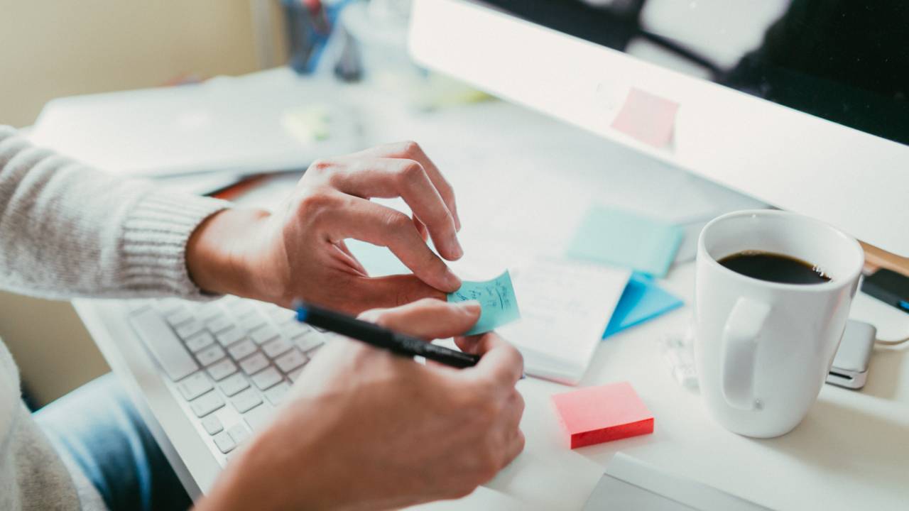 Uma pessoa, sentada em frente a uma mesa com um computador, escreve e cola um post-it