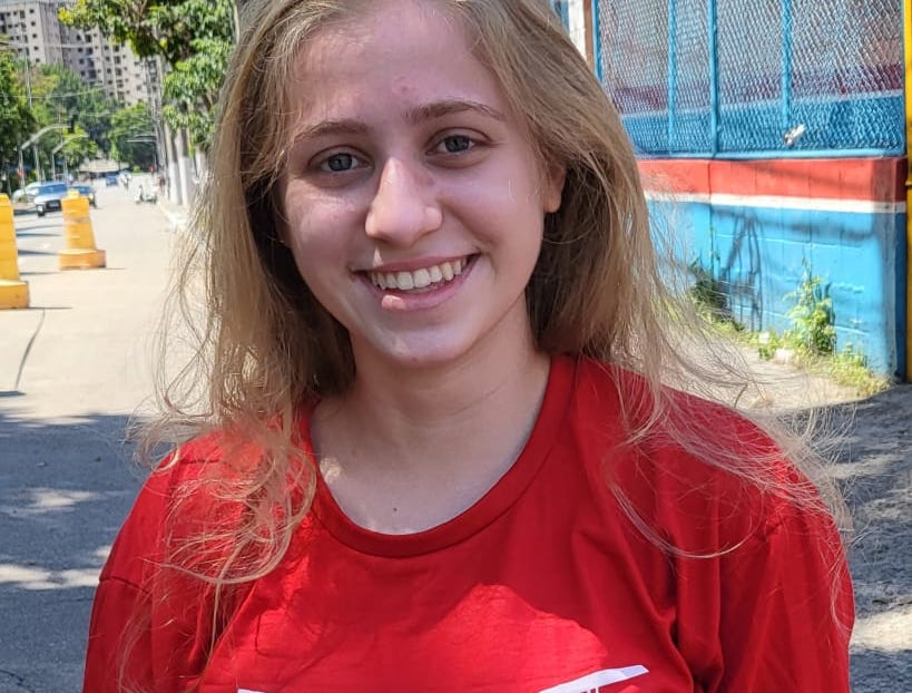 Larissa Mulher jovem branca, loira e de olhos claro, sorri para a câmera usando camiseta vermelha. Ela está na rua.