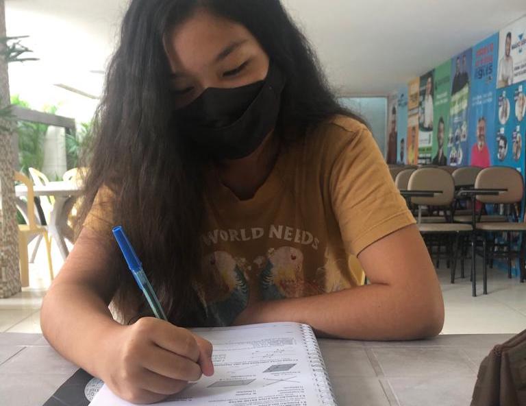 Heloisa Jovem mulher usando máscara de proteção no rosto está sentada escrevendo em um livro. Ela tem pele morena e cabelos negros.