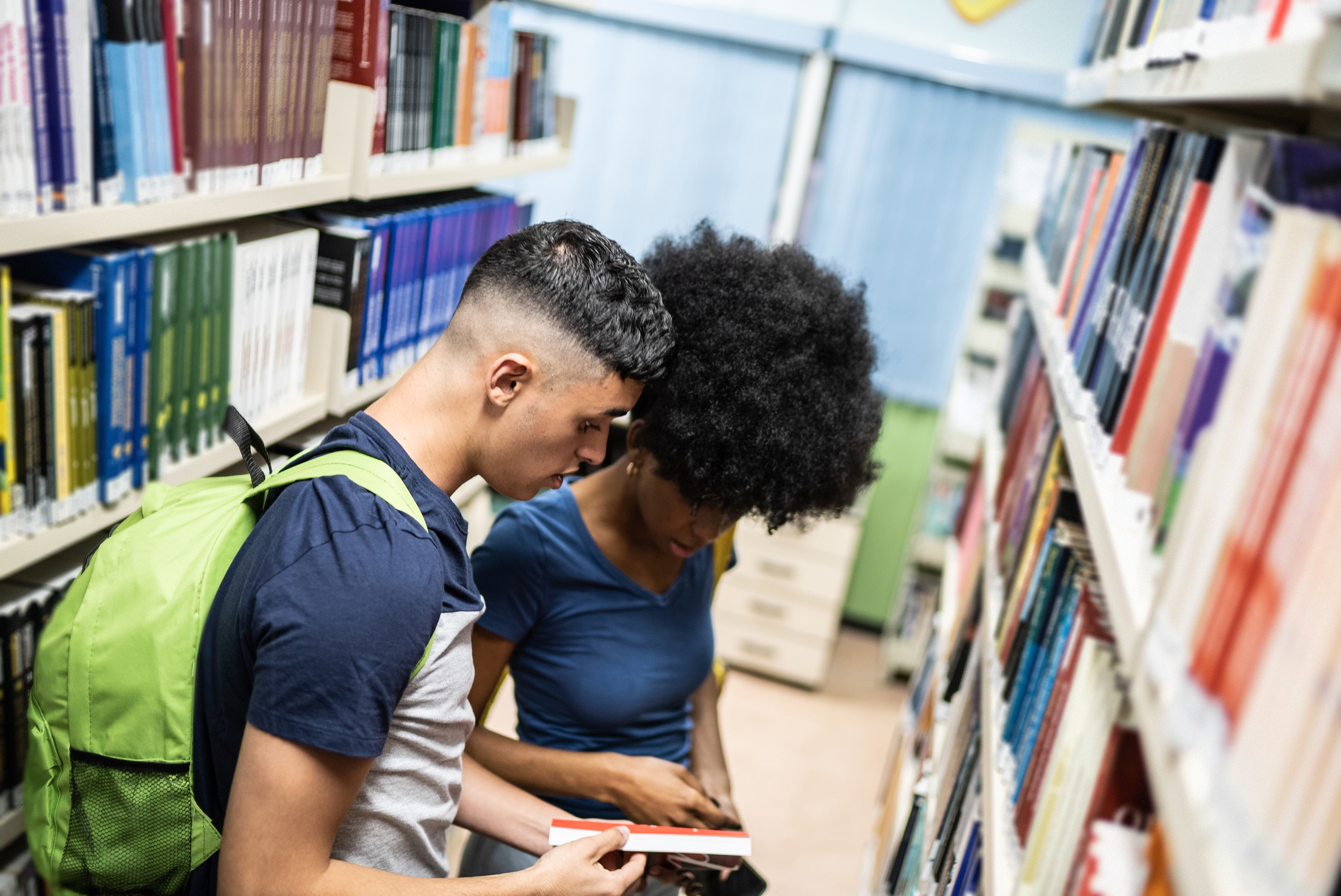 Dois rapazes est&atilde;o folheando um livro em um corredor de uma biblioteca. Um &eacute; branco de cabelo curto e outro &eacute; negro, com cabelo m&eacute;dio