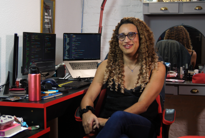 Estudante Nathally Souza sentada em frente a uma mesa, com dois computadores. Ela é negra, usa cabelo trançado e usa óculos de grau.