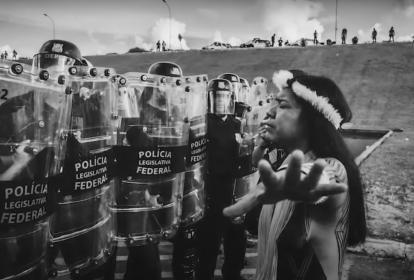 Cena do documentário "Guerras do Brasil.doc" mostra mulher indígena de braços abertos, em frente a batalhão da polícia.