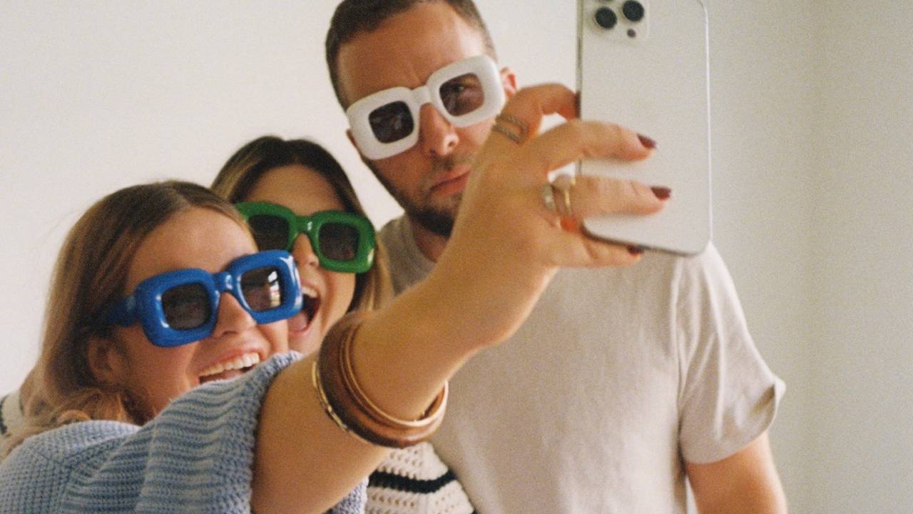 Um homem e duas mulheres vestem óculos de sol com armações coloridas e posam para uma selfie