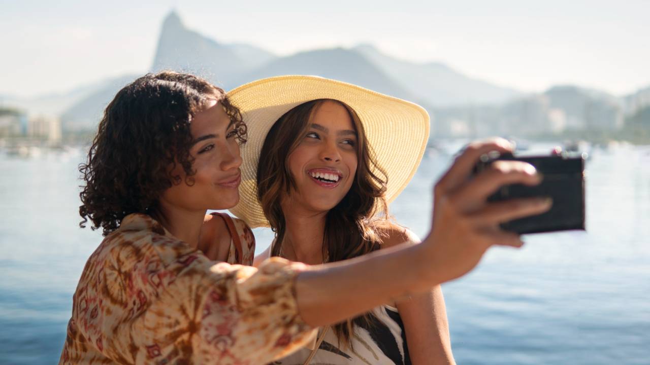 Duas amigas tirando uma selfie em frente a paisagem litorânea, se divertindo, rindo, de férias