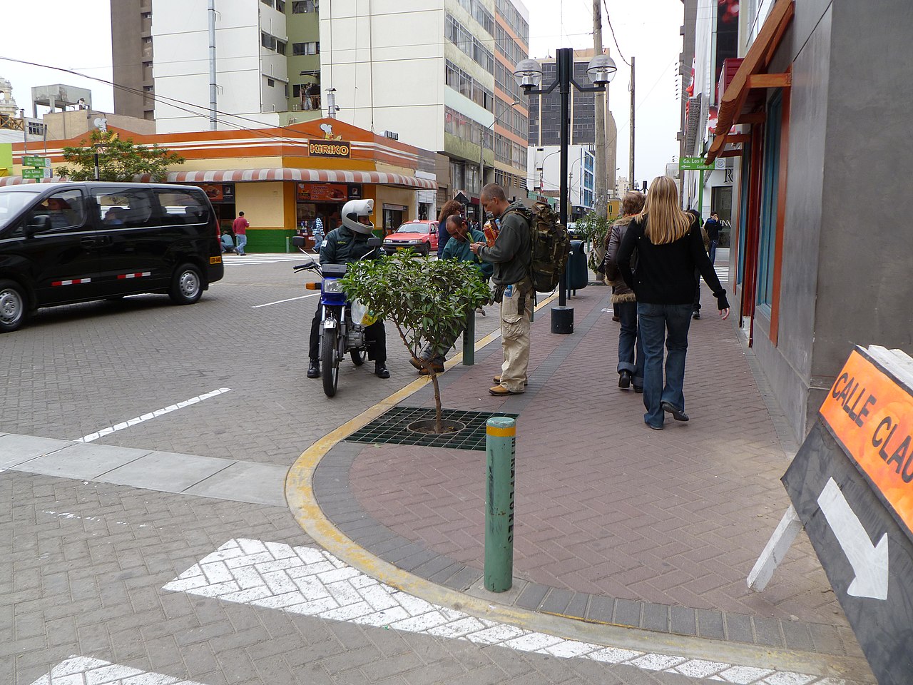 Cena de rua em Lima, Peru, mostrando uma cal&ccedil;ada movimentada com pedestres, um motociclista parado junto a pequenos arbustos em canteiros e edif&iacute;cios comerciais ao fundo em um cruzamento urbano.