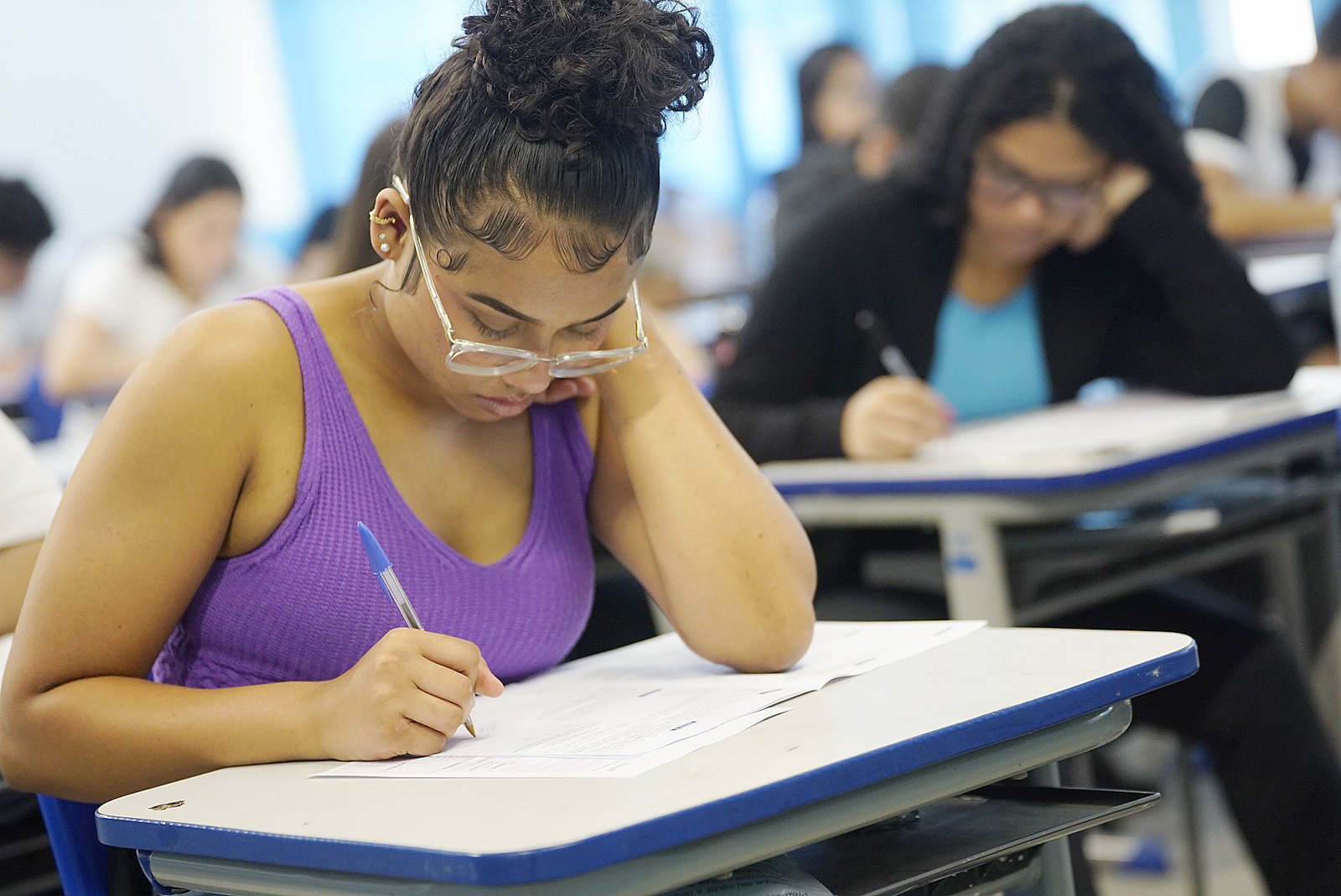 Estudante sentada em carteira escolar, com o rosto desfocado, escrevendo em uma prova com caneta azul em sala de aula cheia de outros alunos também fazendo exame.