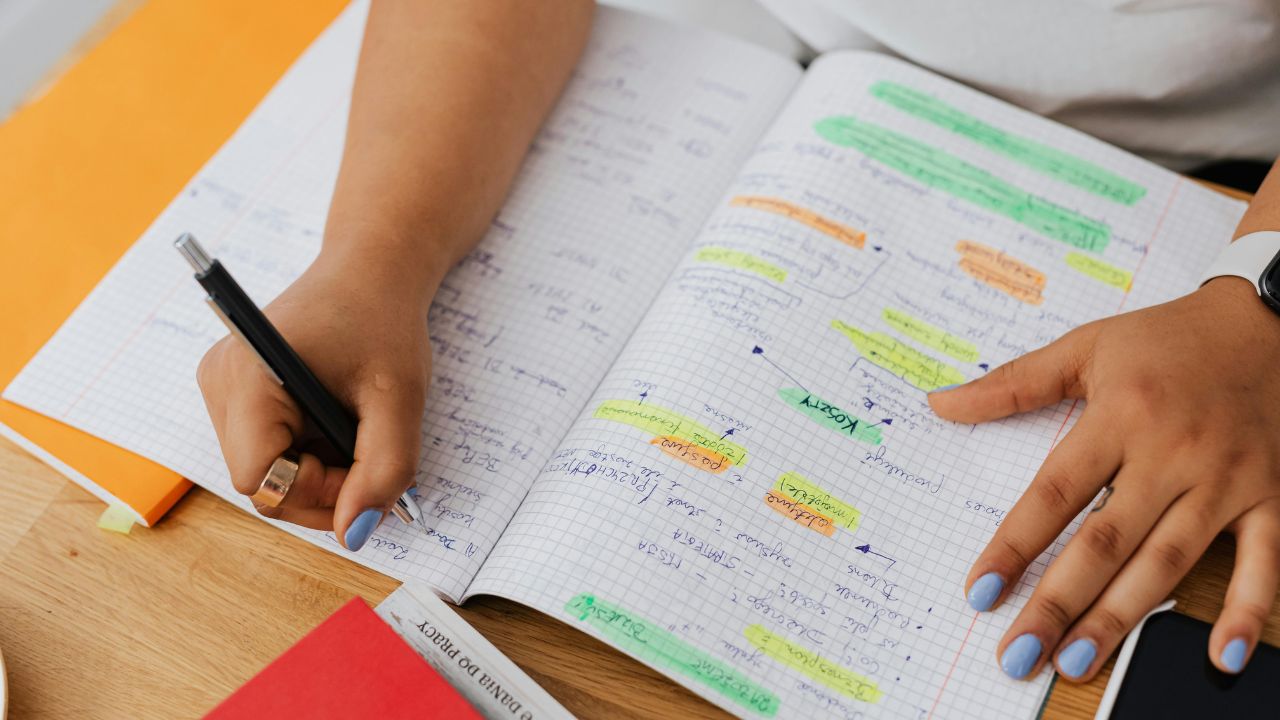 Pessoa registrando em um caderno informações, separadas nas cores laranja, verde e amarelo.