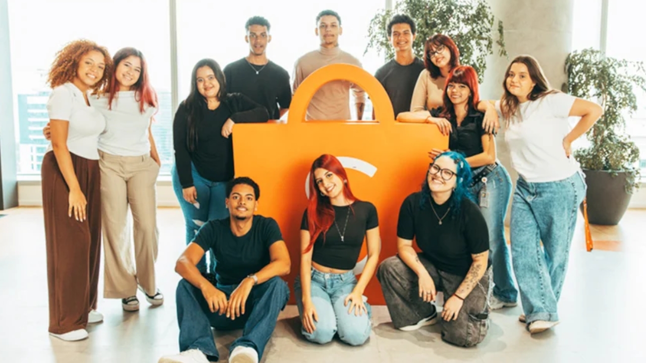 Foto de grupo de 11 jovens diversos (homens e mulheres de diferentes etnias, cabelos coloridos como rosa e azul, tatuagens, roupas casuais modernas como jeans, camisetas brancas e pretas) posando sorridentes ao lado de logotipo gigante laranja da Shopee (sacola de compras estilizada), em escritório moderno com janelas amplas, plantas altas e piso claro, celebrando participação no Programa Jovem Aprendiz