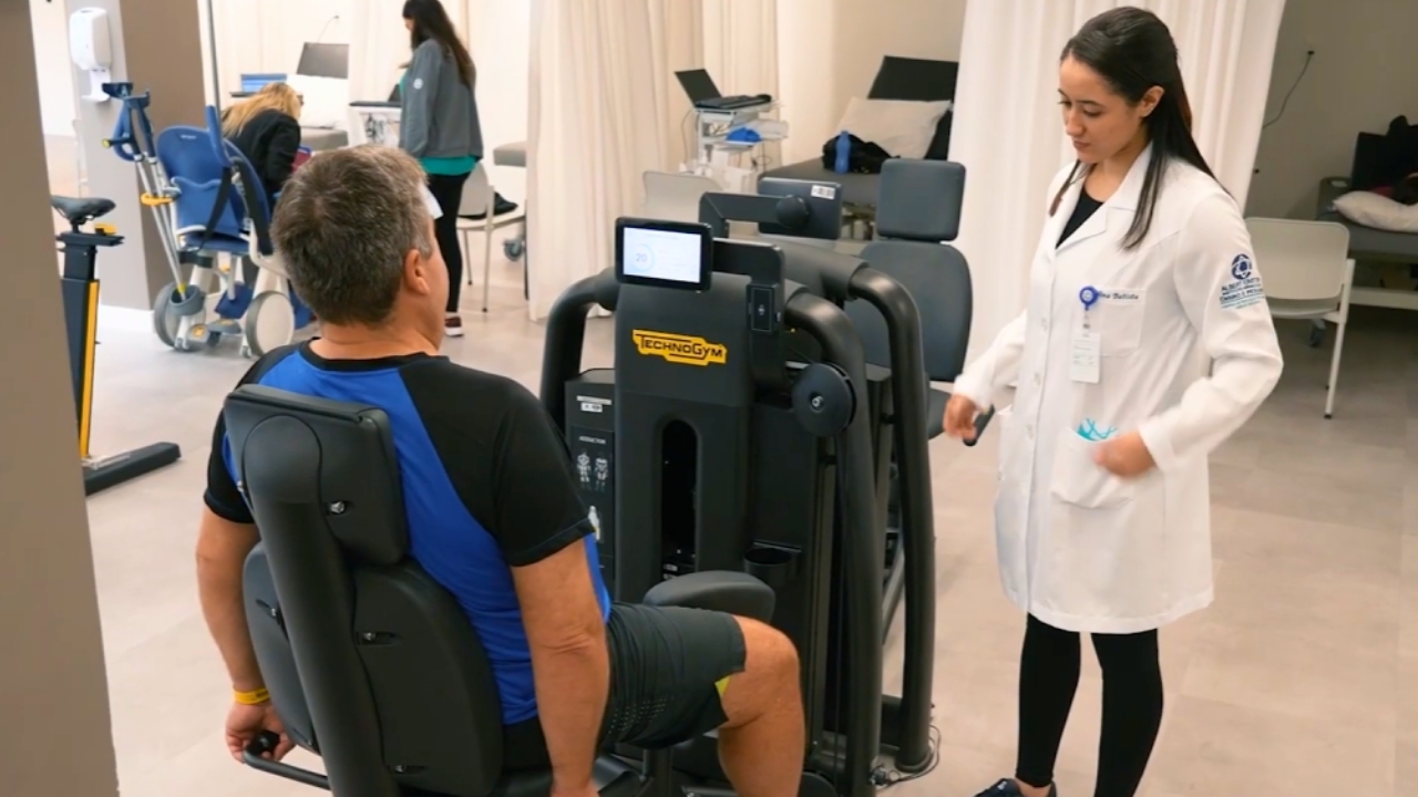 Um homem com roupa de ginástica usa um aparelho de leg press em uma sala de fisioterapia, auxiliado por uma mulher de jaleco branco. A cena transmite uma sensação de reabilitação focada.