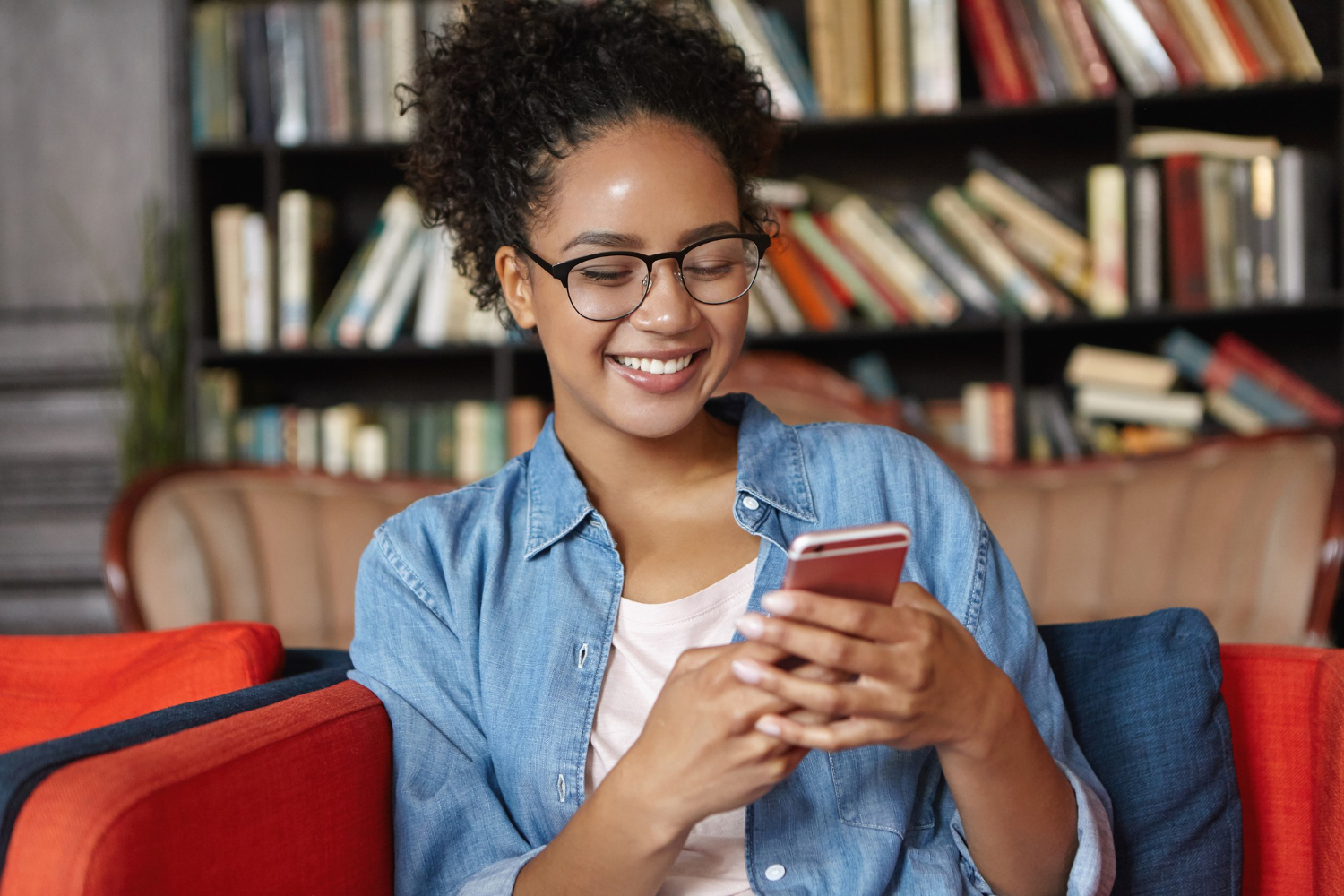 Mulher sorridente de óculos usa smartphone, sentada em uma cadeira colorida em uma biblioteca aconchegante. Livros preenchem as prateleiras ao fundo.