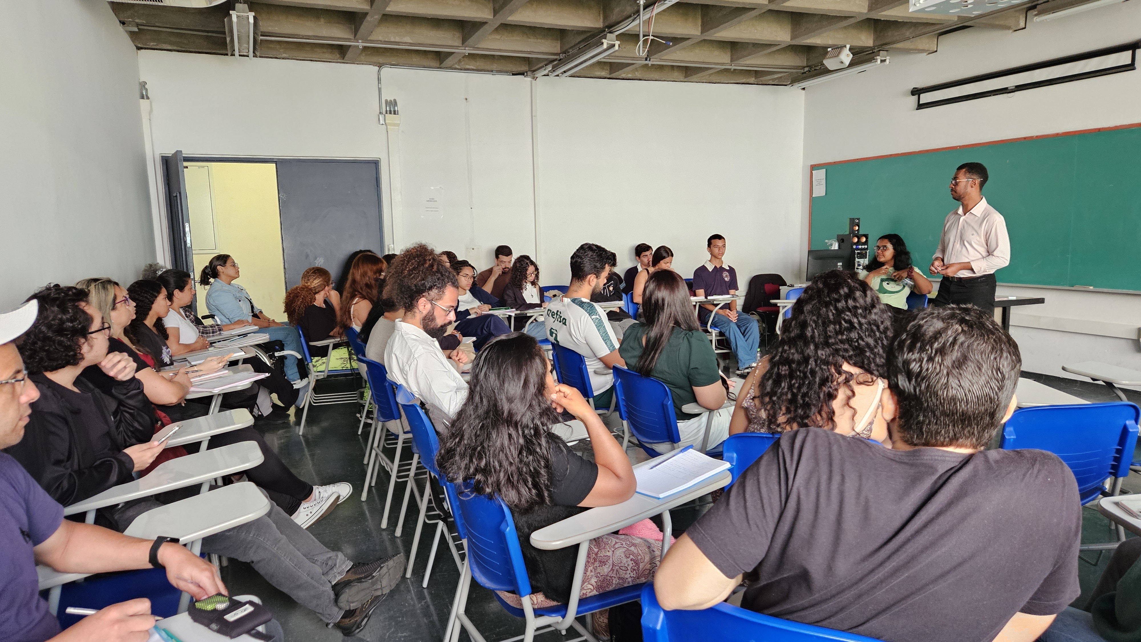 Sala de aula com cerca de 20 alunos sentados em carteiras azuis e brancas, olhando para um professor asiático de camisa clara e calça escura, em pé à direita, perto de um quadro-negro verde. Uma mulher de camiseta verde clara está sentada à mesa do professor, olhando para a turma. O ambiente é iluminado e as paredes são brancas