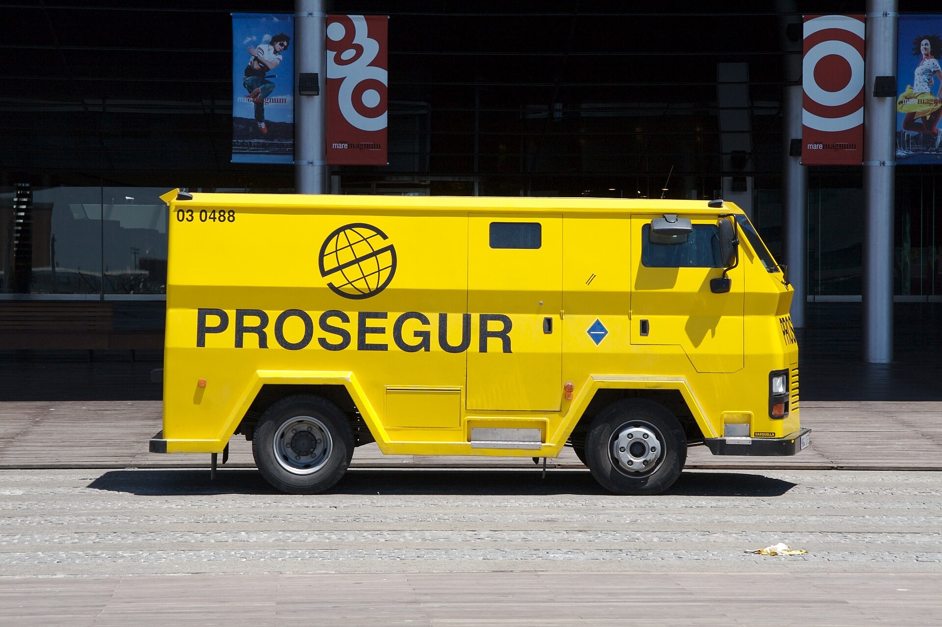 Carro-forte amarelo da Prosegur estacionado, com o número 03 0488 e o logo da empresa, em frente a um prédio moderno com janelas escuras e banners coloridos pendurados