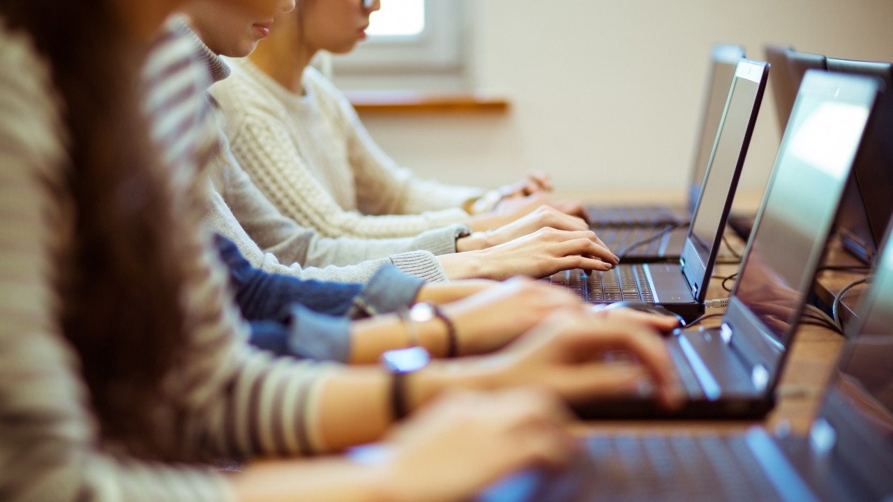 Pessoas sentadas em fila, digitando em notebooks pretos com telas acesas, em um ambiente de sala de aula ou escritório. Mãos focadas nos teclados, indicando concentração e trabalho.