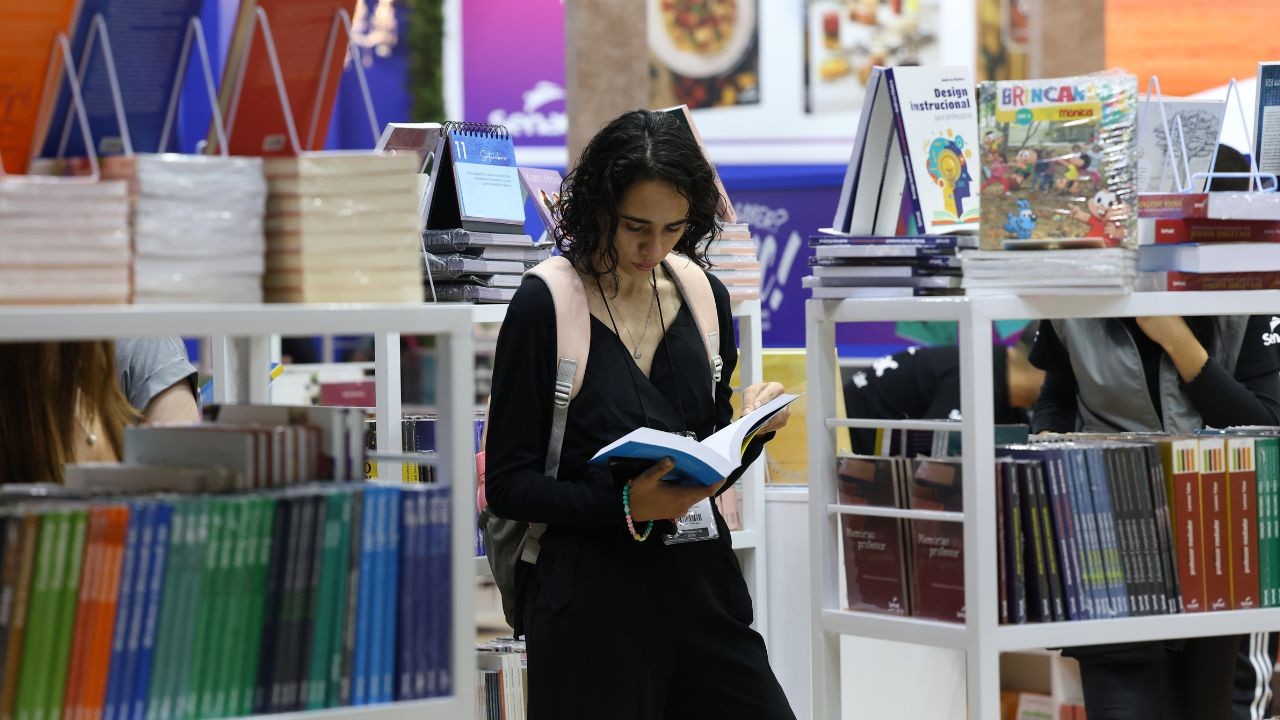 Jovem mulher de cabelos cacheados escuros, vestindo um macacão preto e mochila bege, lendo um livro de capa azul em uma livraria ou feira de livros, com estantes cheias de livros coloridos ao redor