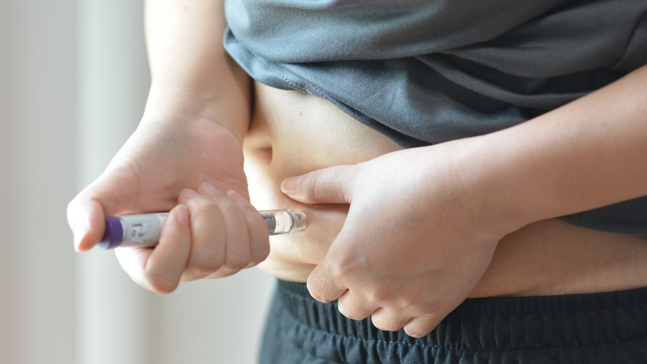Pessoa com camiseta cinza e calça preta aplicando uma injeção na barriga, segurando a pele abdominal com uma mão e o aplicador com a outra